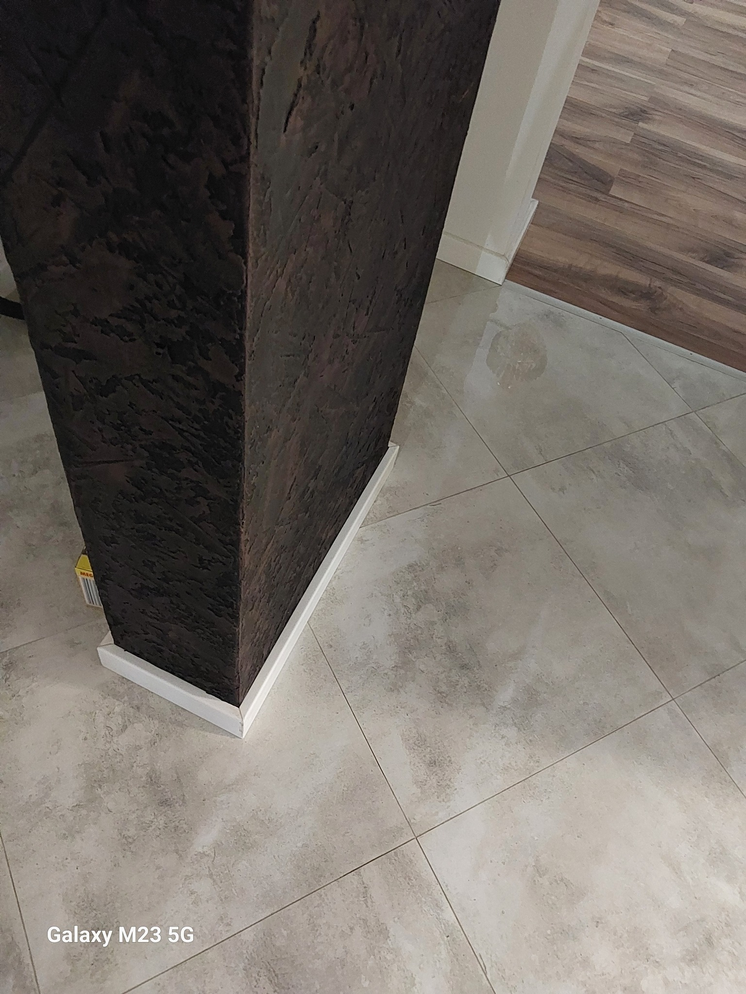 Dekoracyjny filar z ciemną, strukturalną powierzchnią, obudowany białą listwą przypodłogową, na tle szarych płytek podłogowych i fragmentu drewnianej podłogi w przejściu.