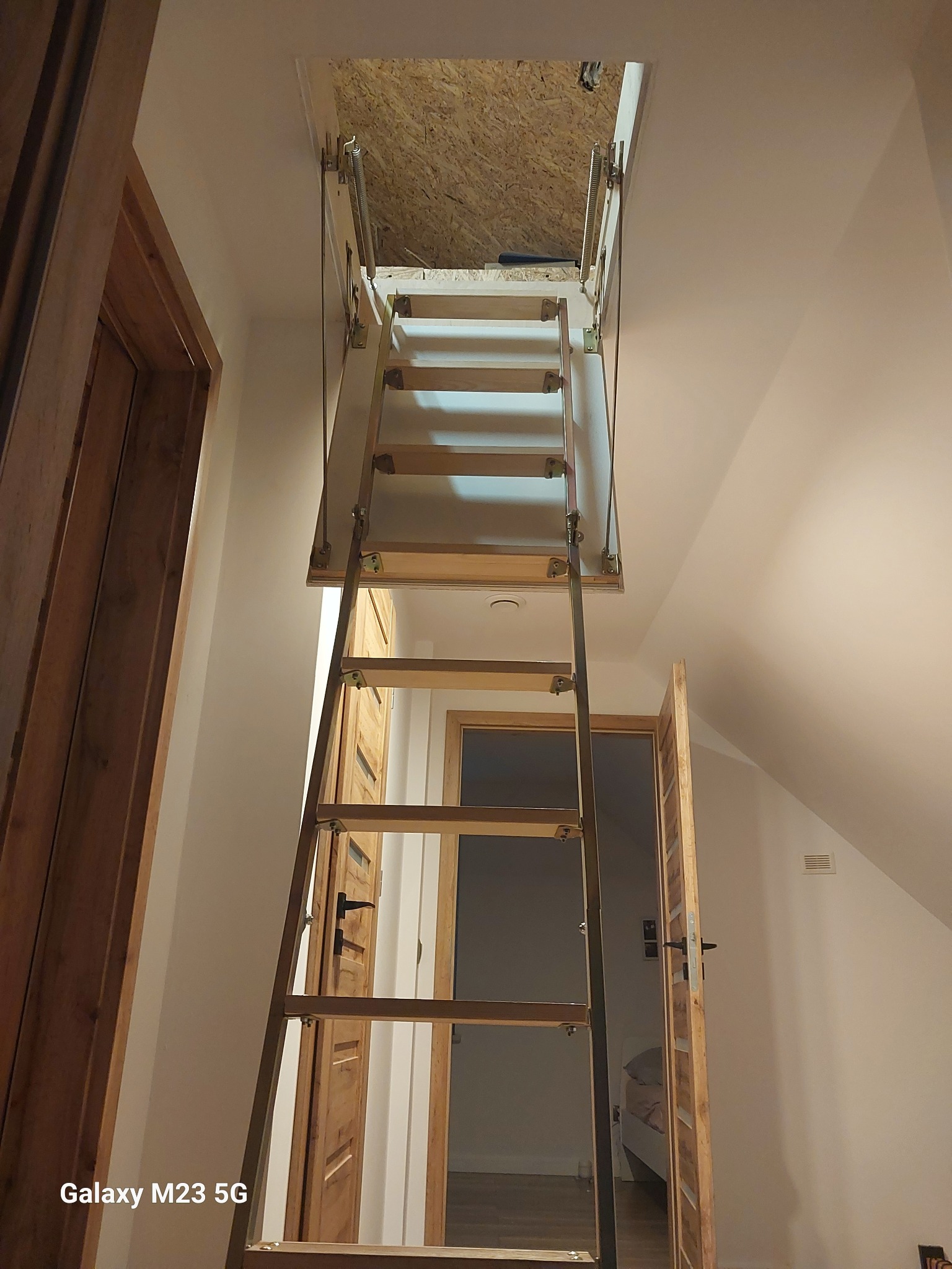 Składane schody strychowe z drewnianymi stopniami, prowadzące do otworu w suficie, widok z dołu na poddasze wyłożone płytą OSB, w tle widoczne drzwi i fragment pokoju na poddaszu.
