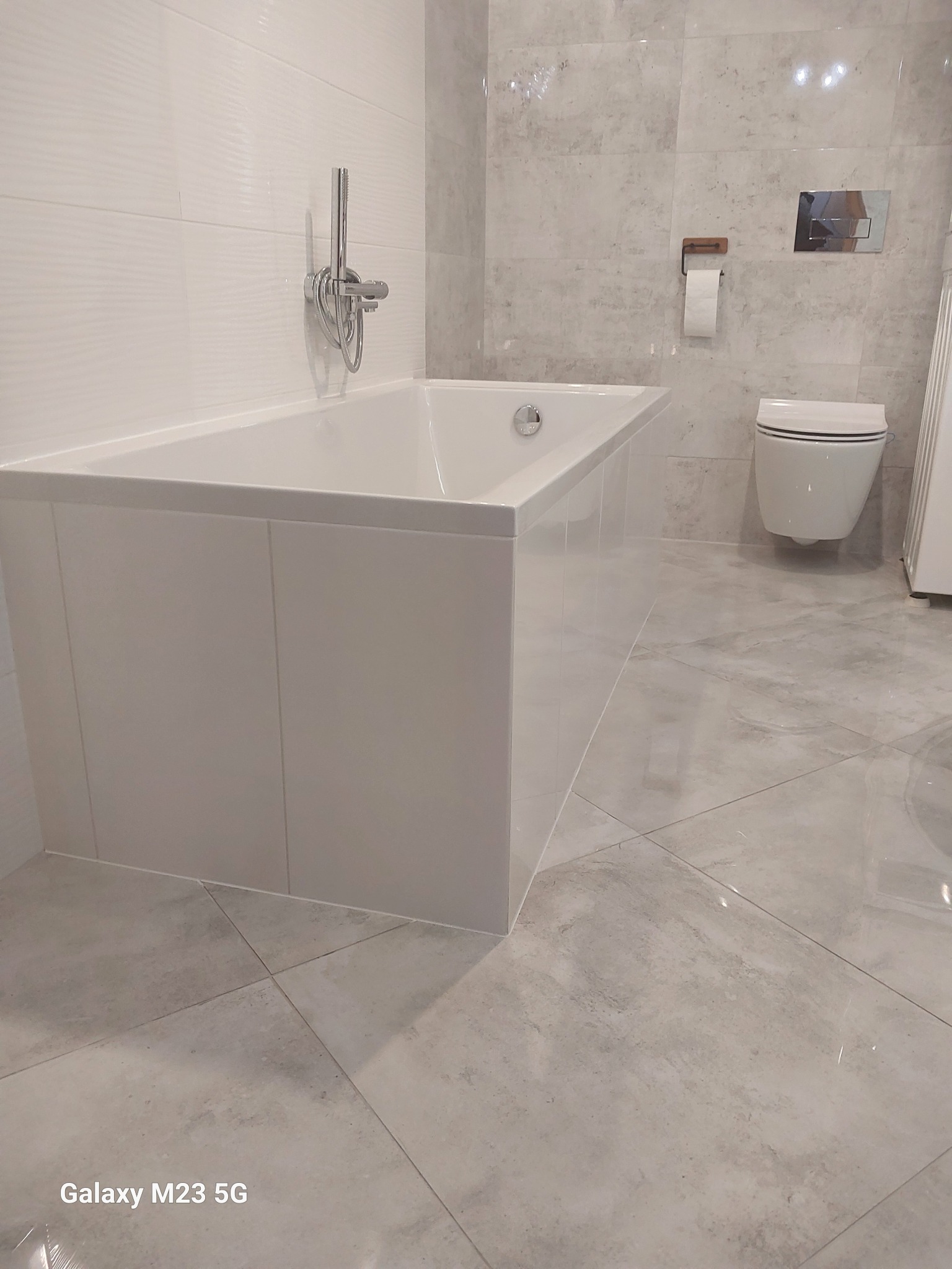 Nowoczesna łazienka z białą wanną obudowaną panelami, chromowaną armaturą, podwieszaną toaletą i szarą marmurkową glazurą na ścianach i podłodze.