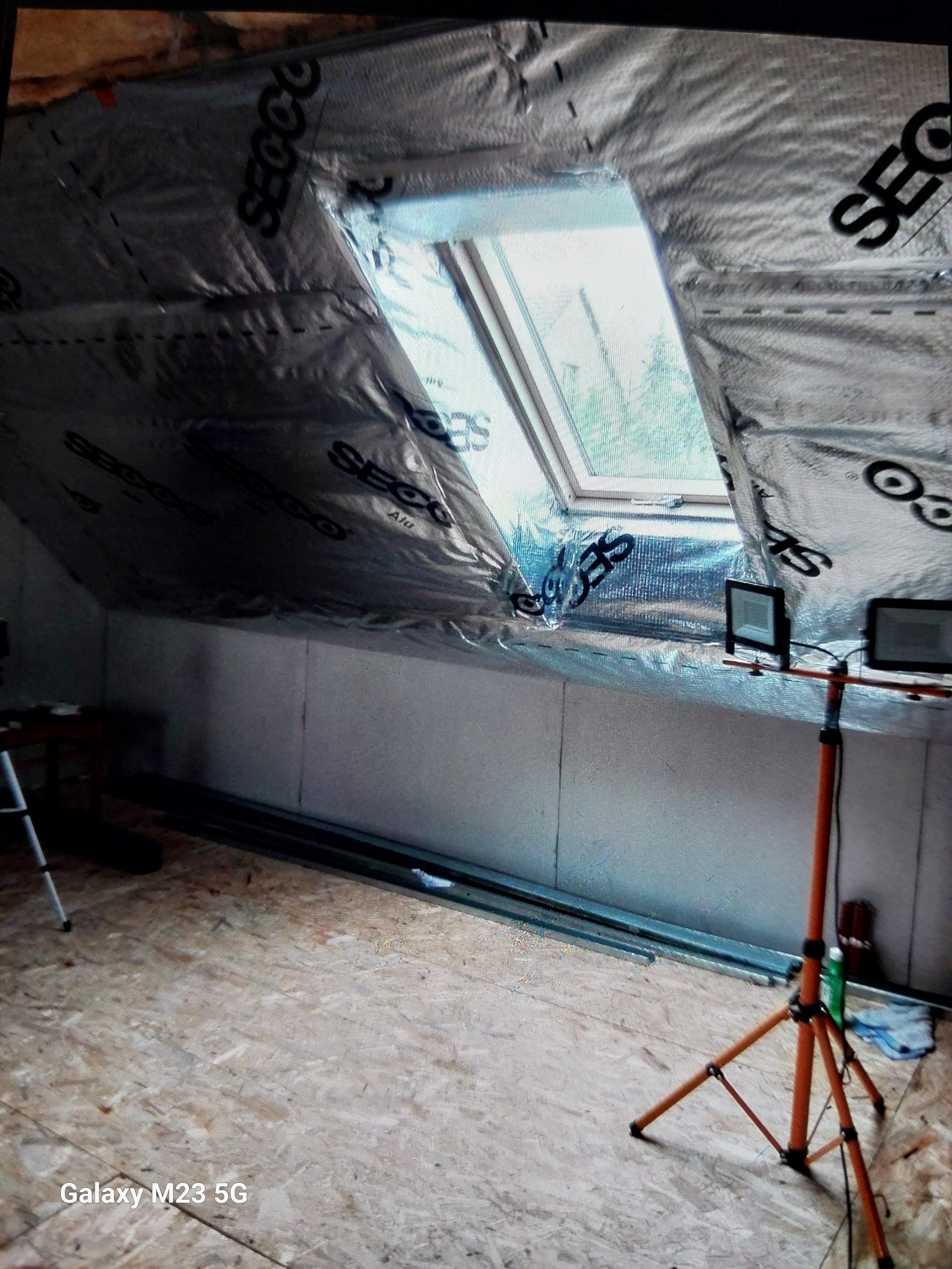 Poddasze w trakcie izolacji, widoczne okno dachowe, folia paroizolacyjna z nadrukiem, stelaż pod płyty gipsowo-kartonowe oraz lampy budowlane na trójnogu.