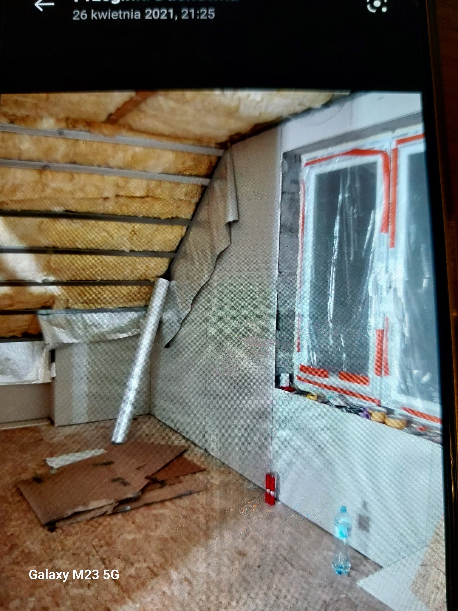 Poddasze w trakcie wykańczania: ocieplenie wełną mineralną, płyty karton-gips na ścianach przy oknie zabezpieczonym folią. Widoczny fragment podłogi i materiały budowlane.