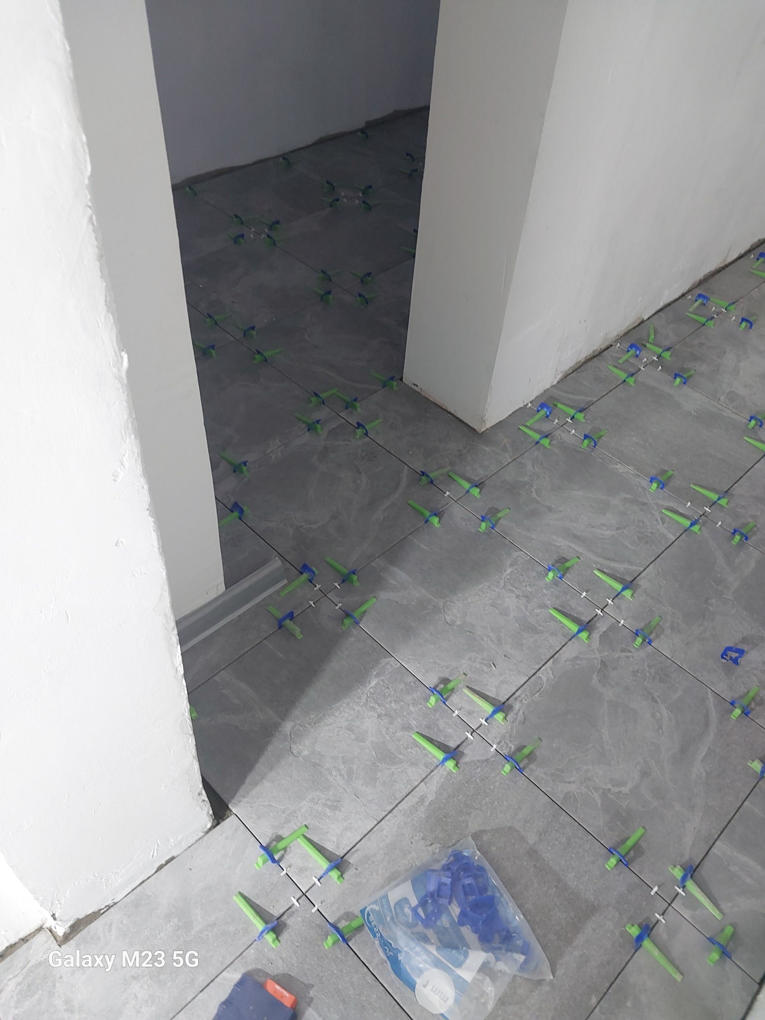 Szare płytki podłogowe w trakcie układania, z użyciem zielonych i niebieskich krzyżyków dystansowych, widoczne narzędzia i woreczek z klinami.