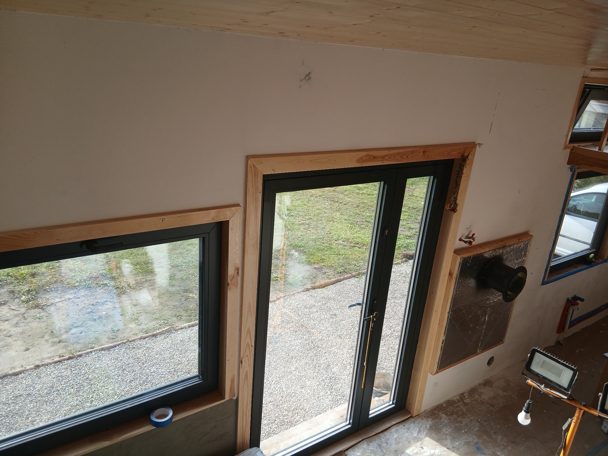 Wnętrze w trakcie wykańczania: okna z drewnianą ramą, drzwi balkonowe, fragment ocieplenia ściany z wentylacją, sufit z boazerii, lampa budowlana. Widok z góry.