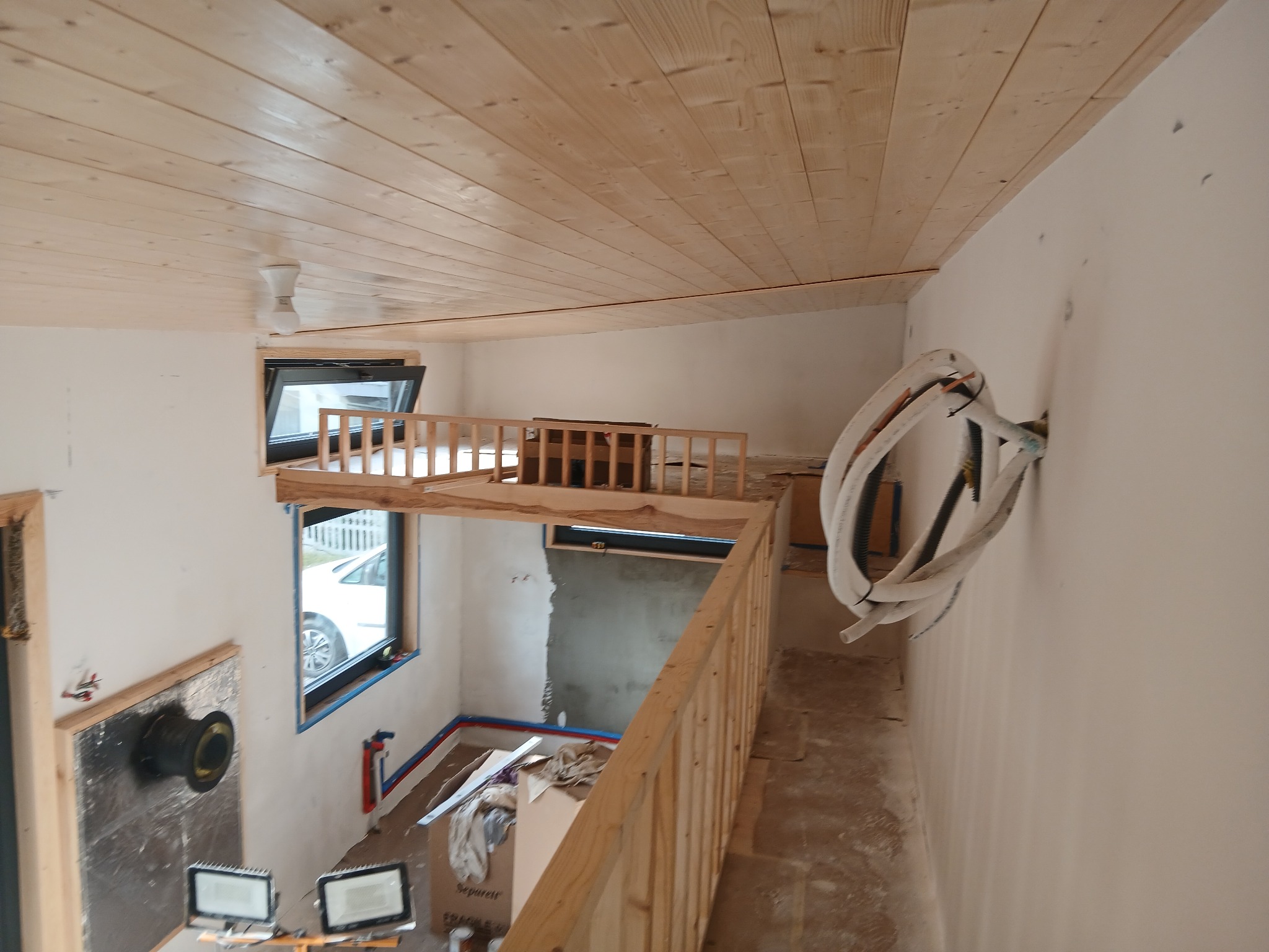 Wnętrze domu w trakcie wykańczania: drewniana boazeria na suficie, balustrada antresoli, instalacje na ścianie, okna. Widoczne materiały budowlane i narzędzia.
