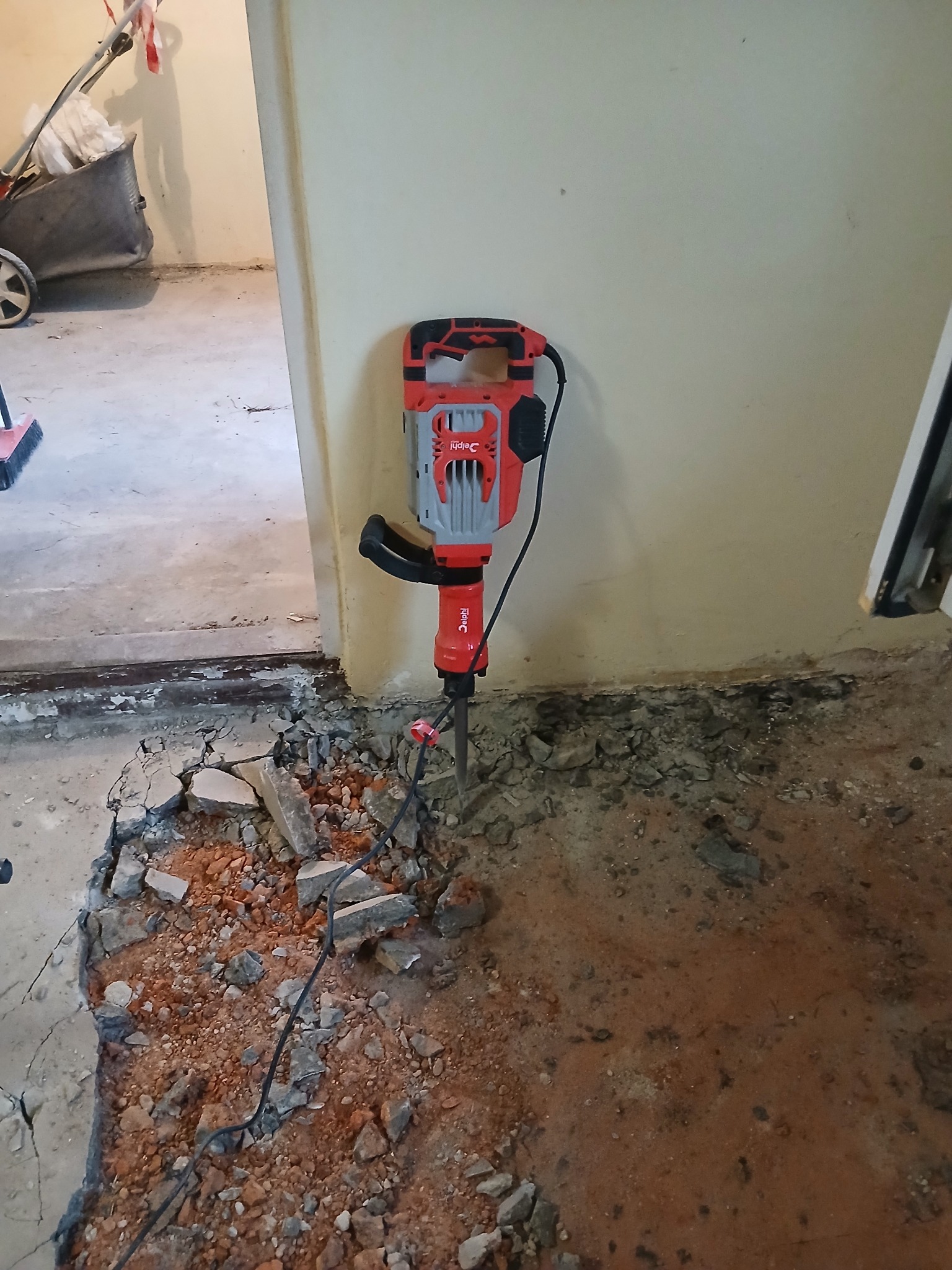 Czerwono-szary młot udarowy wbity w gruz na podłodze podczas skuwania posadzki w pomieszczeniu. Widoczne fragmenty ściany i sprzęt budowlany w tle.