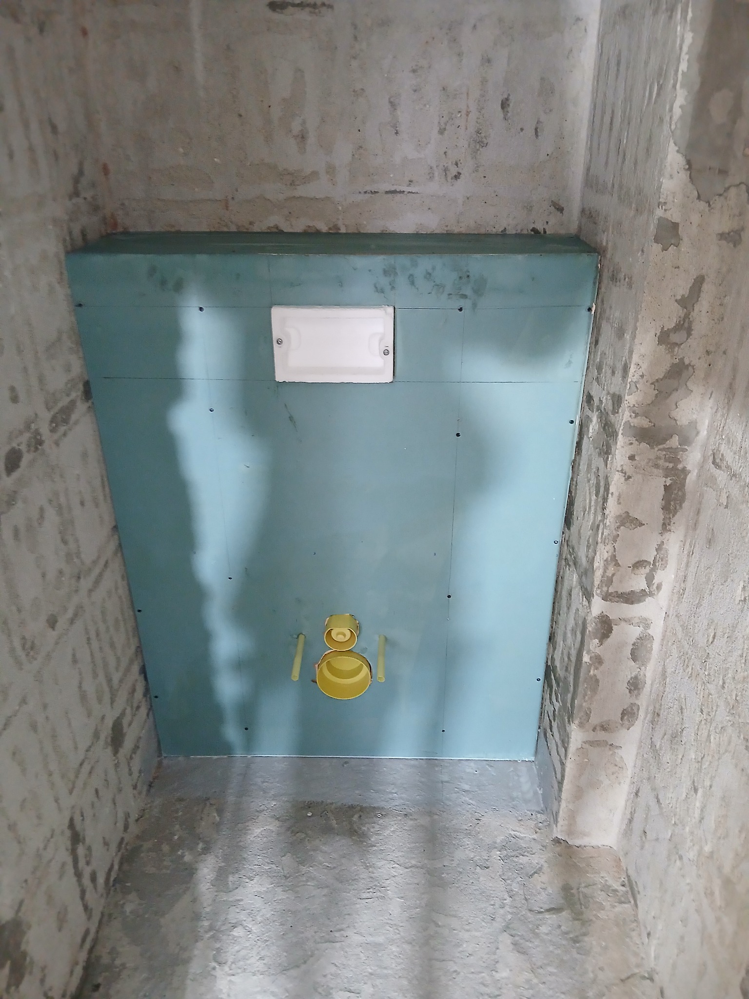 Niebieska płyta karton-gips zamontowana na ścianie w surowej łazience, widoczne żółte elementy instalacji. Ściany i podłoga w stanie deweloperskim.