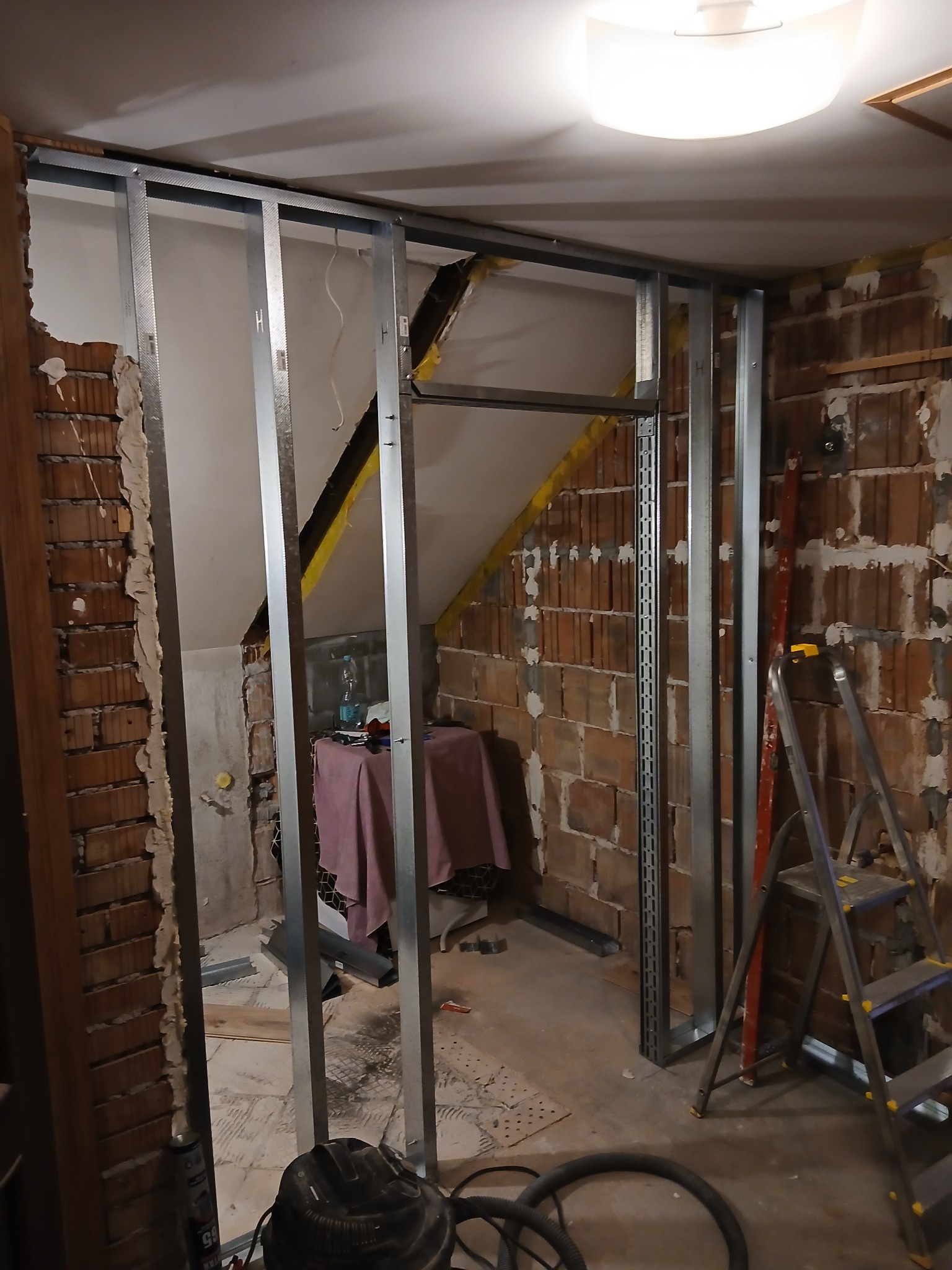 Konstrukcja ścianki działowej z profili stalowych na tle surowej cegły i fragmentu poddasza. Widoczne narzędzia i drabina. Etap prac wykończeniowych.