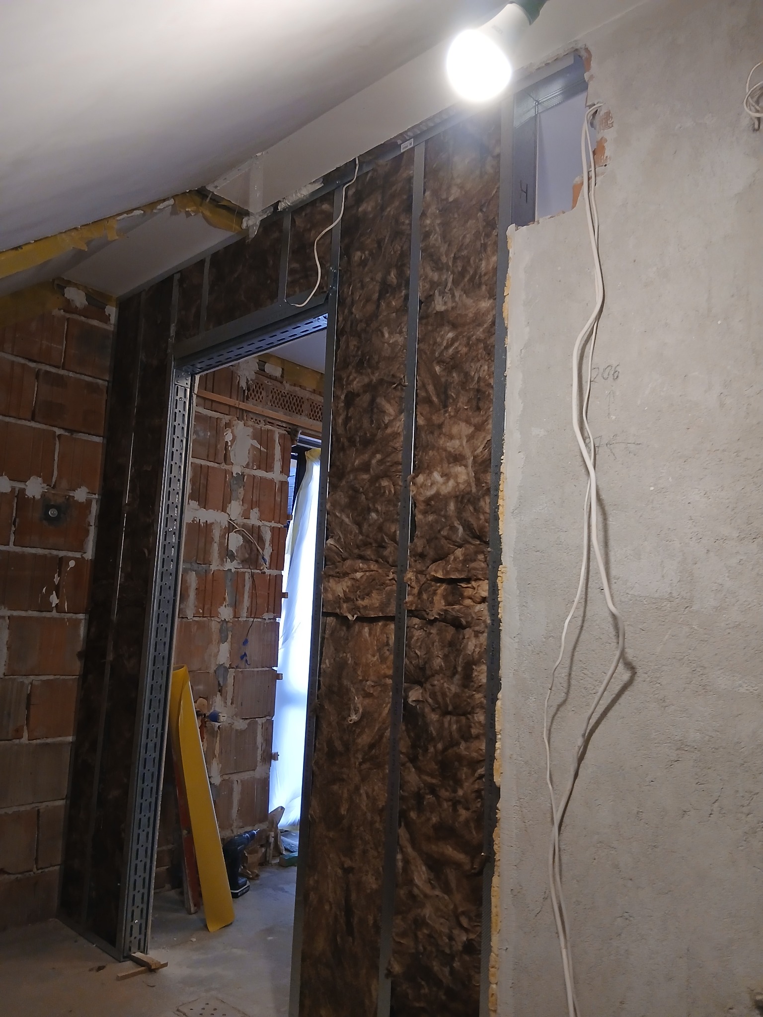 Montaż izolacji z wełny mineralnej w metalowym stelażu, widoczne przewody elektryczne na surowej ścianie. Etap prac wykończeniowych wewnątrz budynku.