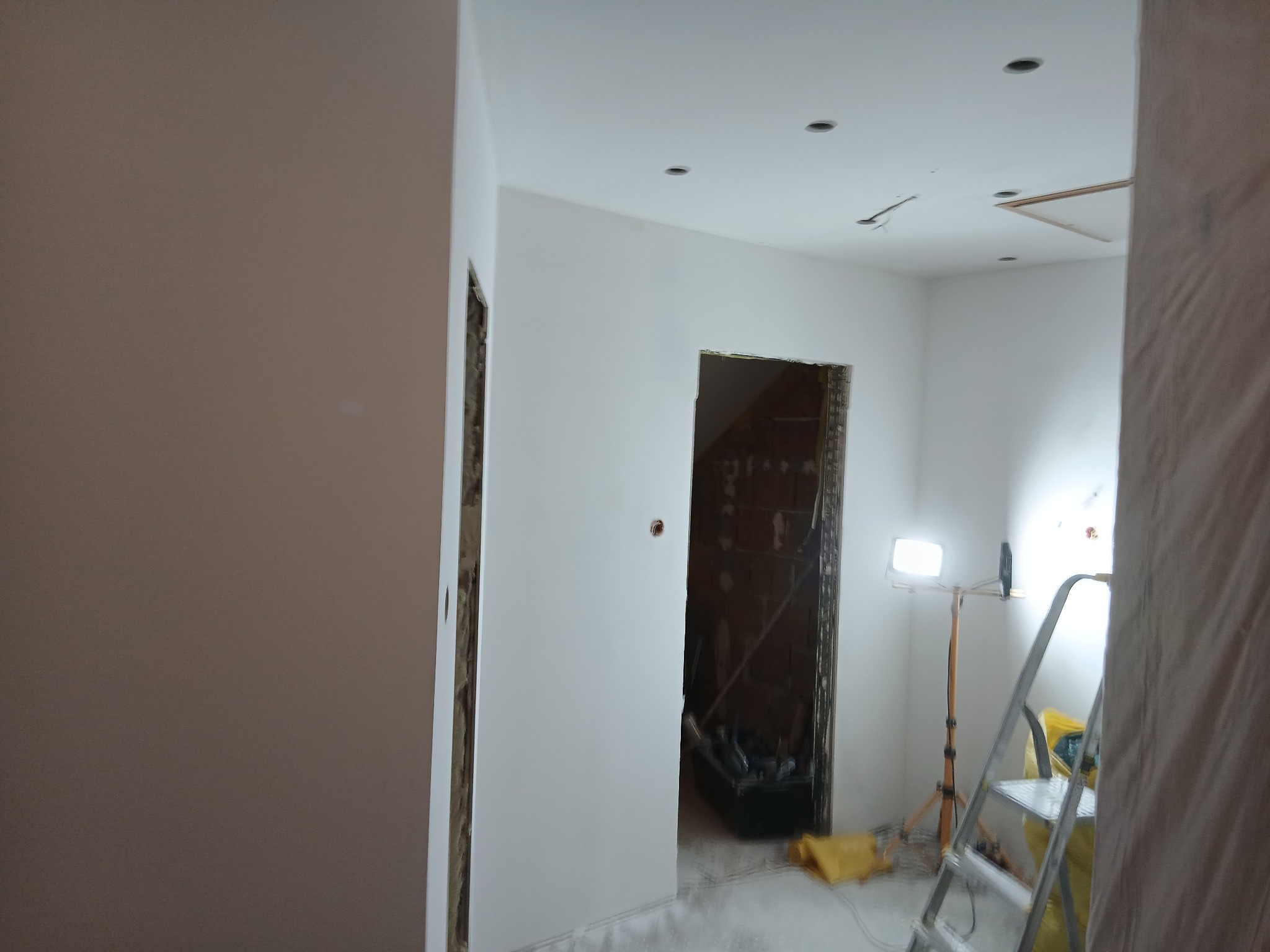 Wnętrze w trakcie remontu: świeżo pomalowane ściany na biało, widoczne otwory drzwiowe w stanie surowym, drabina i lampa budowlana. Etap prac wykończeniowych.