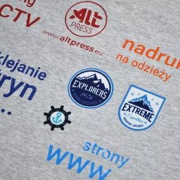 Alt-Press Krzysztof Żółtowski - Szara koszulka z różnorodnymi nadrukami: logo firmy ALT Press, hasła reklamowe dotyczące oklejania witryn, tworzenia stron WWW, nadruków na odzieży, monitoringu CCTV oraz grafiki związane...