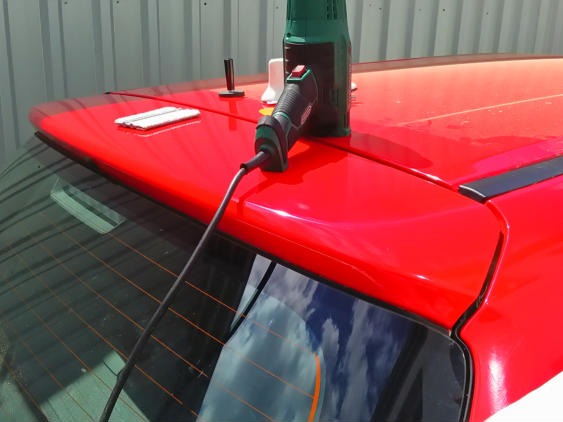 Aplikacja folii na tylną szybę czerwonego samochodu za pomocą opalarki, widoczne narzędzia i fragmenty folii.