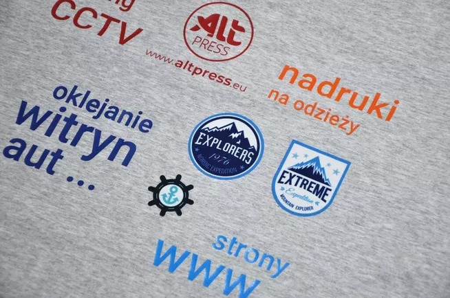 Szara koszulka z różnorodnymi nadrukami: logo firmy ALT Press, hasła reklamowe dotyczące oklejania witryn, tworzenia stron WWW, nadruków na odzieży, monitoringu CCTV oraz grafiki związane...