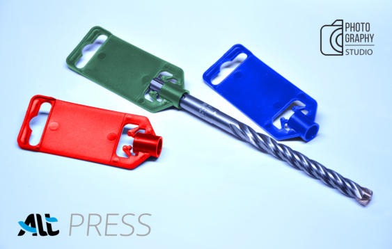 Wiertło do betonu z trzema kolorowymi, plastikowymi zawieszkami: czerwona, zielona i niebieska, leżące na białym tle. Widoczne logo firmy fotograficznej.