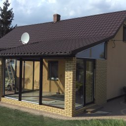 U. Kozioł, G. Kozioł sp. jw. - Nowoczesna oranżeria z cegły klinkierowej, z dużymi przeszkleniami i brązowym dachem, widoczna drabina oparta o ścianę i basen ogrodowy w tle.