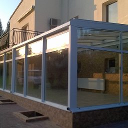 U. Kozioł, G. Kozioł sp. jw. - Zewnętrzny widok przeszklonej, nowoczesnej werandy z białymi ramami, przylegającej do budynku z balkonem i klimatyzacją, otoczonej zielenią i kostką brukową.