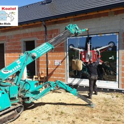 U. Kozioł, G. Kozioł sp. jw. - Montaż okna na budowie domu jednorodzinnego za pomocą specjalistycznego miniżurawia Maeda; pracownik w czarnej odzieży kontroluje proces wstawiania szyby z przyssawkami, widoczne logo firmy Okna...