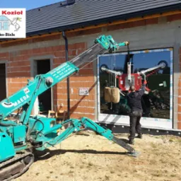 Montaż okna na budowie domu jednorodzinnego za pomocą specjalistycznego miniżurawia Maeda; pracownik w czarnej odzieży kontroluje proces wstawiania szyby z przyssawkami, widoczne logo firmy Okna...