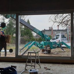 U. Kozioł, G. Kozioł sp. jw. - Montaż okna panoramicznego z użyciem przyssawkowej windy i miniżurawia gąsienicowego; widoczni dwaj monterzy, narzędzia oraz transport okien na przyczepie.