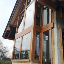 U. Kozioł, G. Kozioł sp. jw. - Nowoczesna fasada domu w trakcie budowy z zamontowanymi oknami z drewnopodobnymi ramami, widoczne elementy konstrukcyjne i ocieplenie.