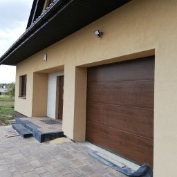 U. Kozioł, G. Kozioł sp. jw. - Nowoczesna brama garażowa segmentowa w kolorze ciemnego drewna, zamontowana w elewacji domu o beżowym tynku, z widocznymi schodami wejściowymi i kostką brukową.