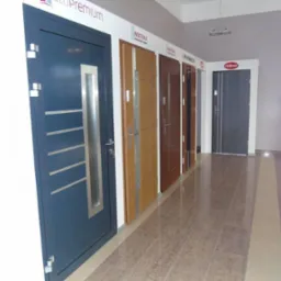 Ekspozycja różnych modeli drzwi zewnętrznych w salonie, widoczne drzwi antywłamaniowe z przeszkleniem i metalowymi listwami oraz inne modele z różnymi wykończeniami.
