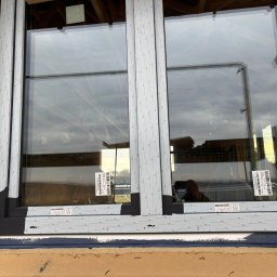 U. Kozioł, G. Kozioł sp. jw. - Montaż dwuskrzydłowego okna z czarną ramą, zabezpieczonego taśmą montażową z nadrukiem, widoczne etykiety z kodami kreskowymi na szybach, oraz żółta taśma zabezpieczająca na górnym rogu ramy.