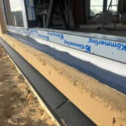 Widok z bliska na montaż okna, widoczna warstwa izolacyjna z pianki, listwa okienna z logo Kömmmerling i świeżo pomalowana rama okienna w kolorze ciemnoniebieskim.