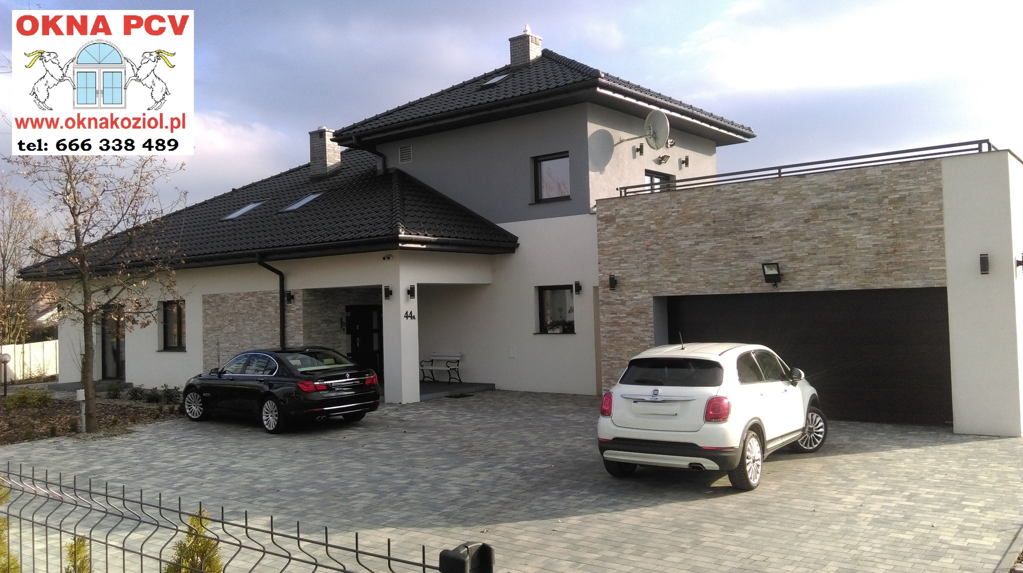 Nowoczesny dom jednorodzinny z czarną bramą garażową, oknami i dachem, na podjeździe stoją dwa samochody, widoczny zza ogrodzenia.