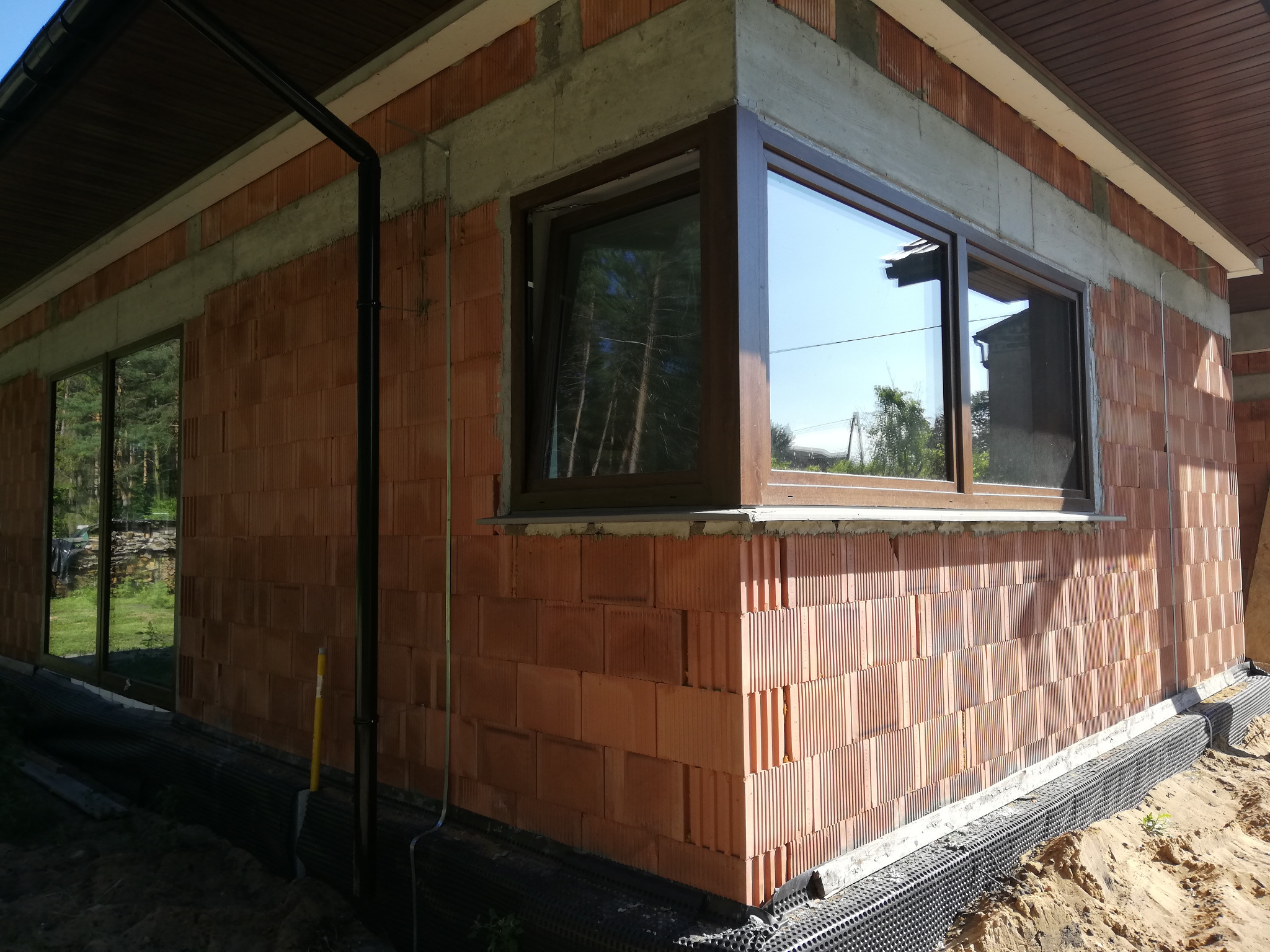 Surowy budynek z cegły ceramicznej z zamontowanymi brązowymi oknami z widocznym odbiciem drzew i nieba, fundament zabezpieczony folią kubełkową.