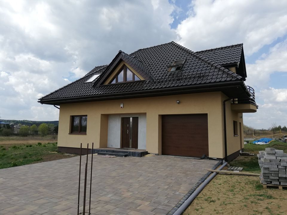 Nowoczesny dom jednorodzinny z brązową bramą garażową, jasną elewacją i brukowanym podjazdem w trakcie wykańczania.