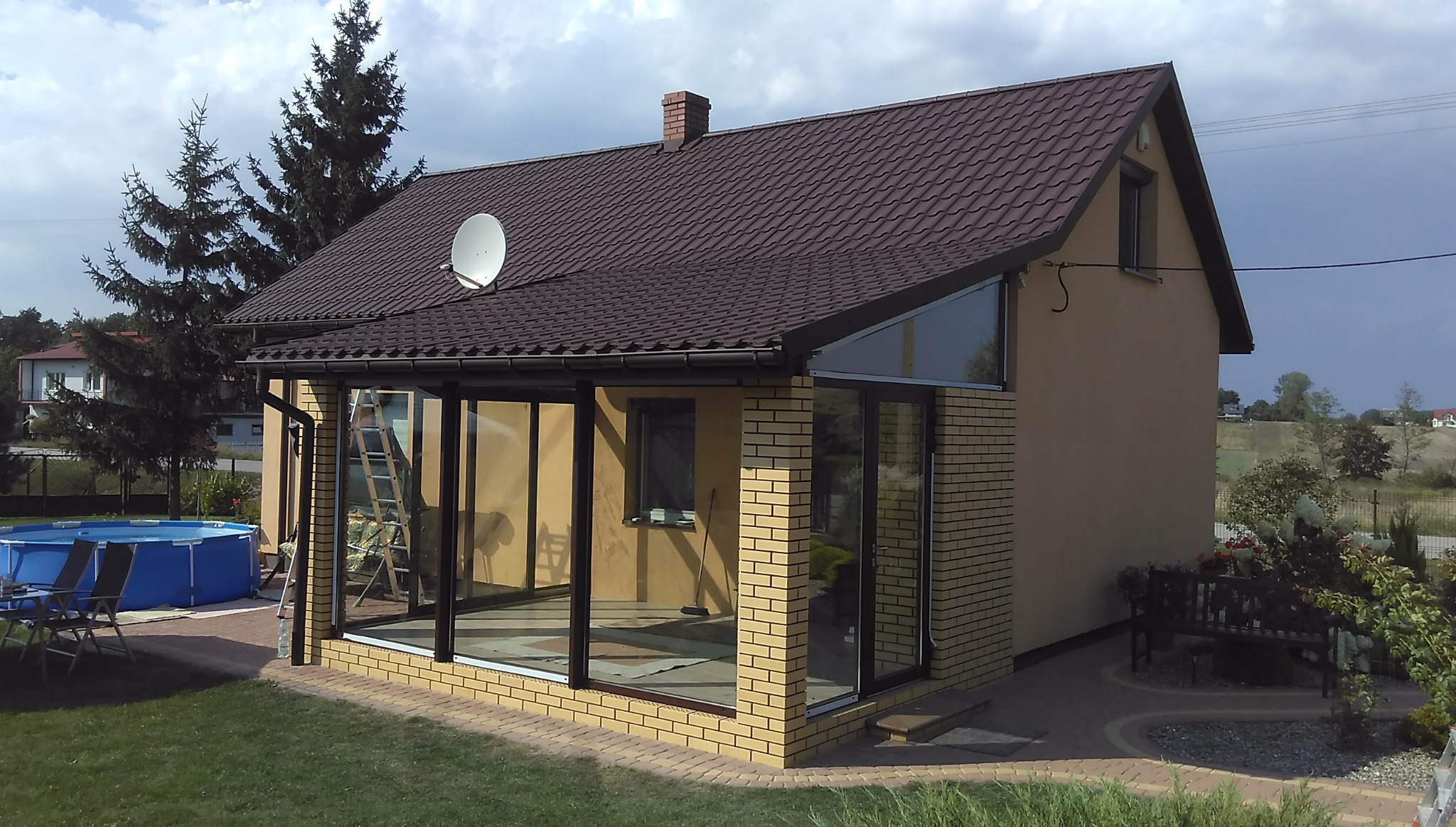 Nowoczesna oranżeria z cegły klinkierowej, z dużymi przeszkleniami i brązowym dachem, widoczna drabina oparta o ścianę i basen ogrodowy w tle.