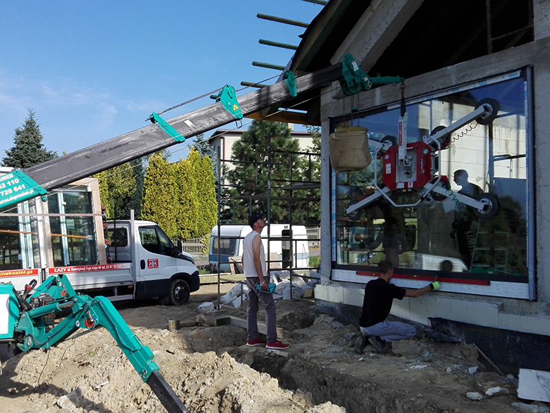 Montaż okna z użyciem podnośnika próżniowego, pracownicy poziomują okno, widoczny samochód dostawczy z oknami w tle.