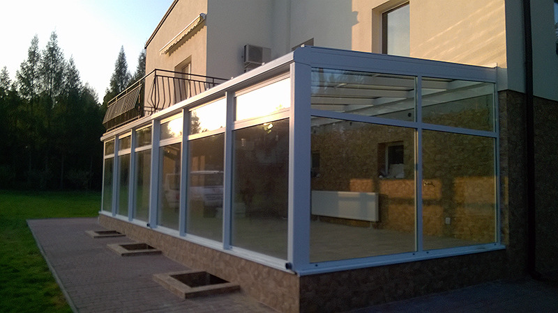 Zewnętrzny widok przeszklonej, nowoczesnej werandy z białymi ramami, przylegającej do budynku z balkonem i klimatyzacją, otoczonej zielenią i kostką brukową.