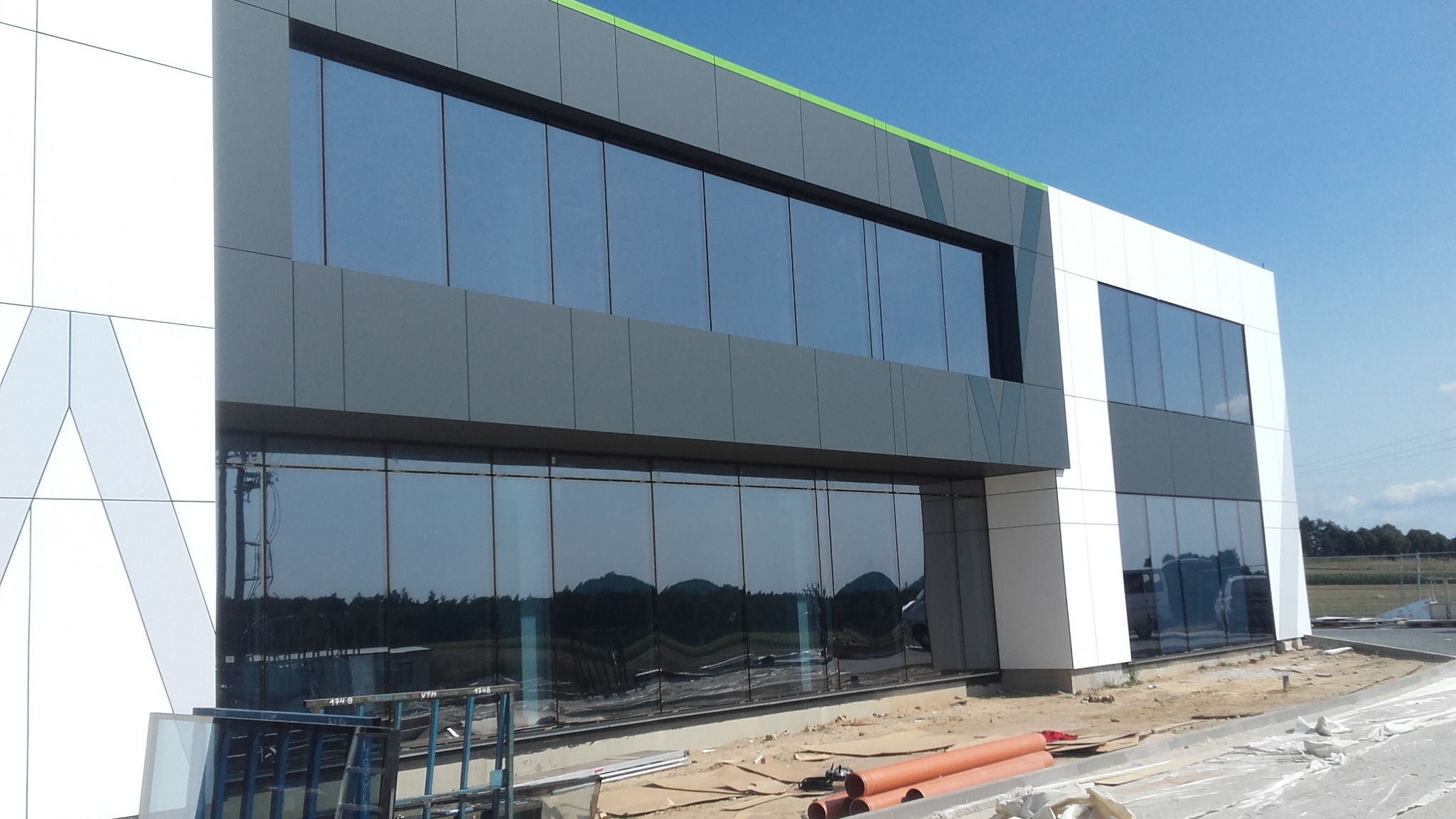 Nowoczesny budynek biurowy z fasadą z paneli aluminiowych i dużymi oknami, widoczne elementy montażowe i rury na ziemi.