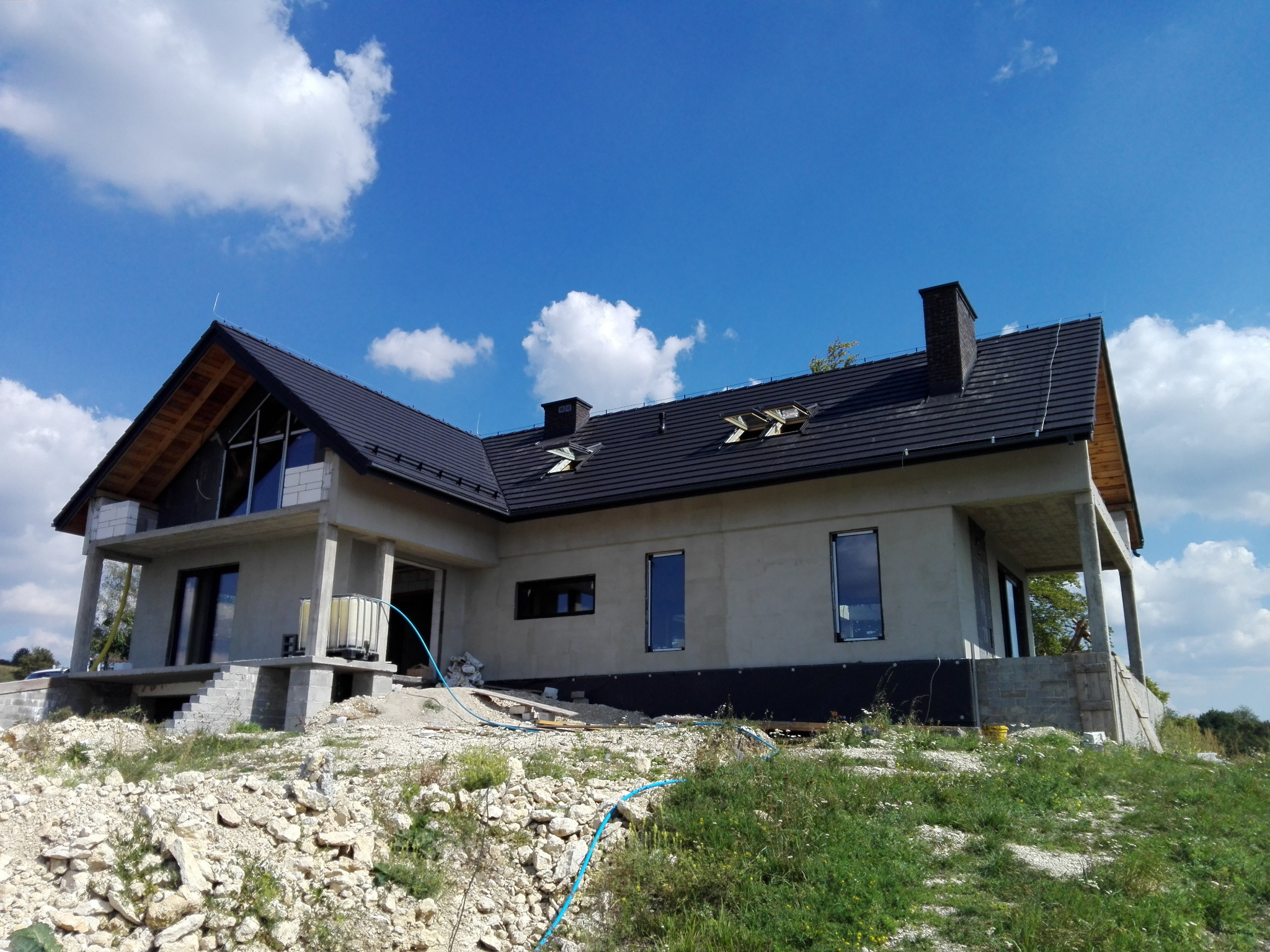 Surowy stan domu jednorodzinnego z czarną dachówką, widoczne okna wstawione, elewacja w trakcie prac, teren wokół domu nierówny, z gruzem i roślinnością.