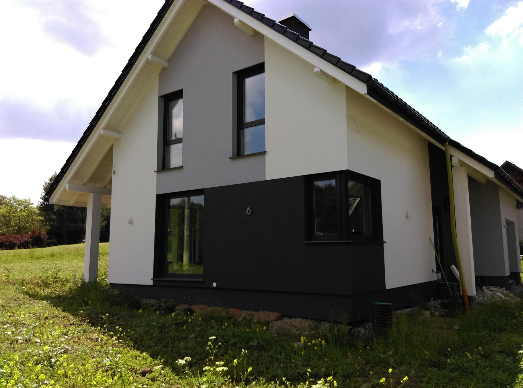 Nowoczesny dom jednorodzinny z czarnymi oknami i elementami elewacji, otoczony zielonym trawnikiem z kwiatami, widok z boku.