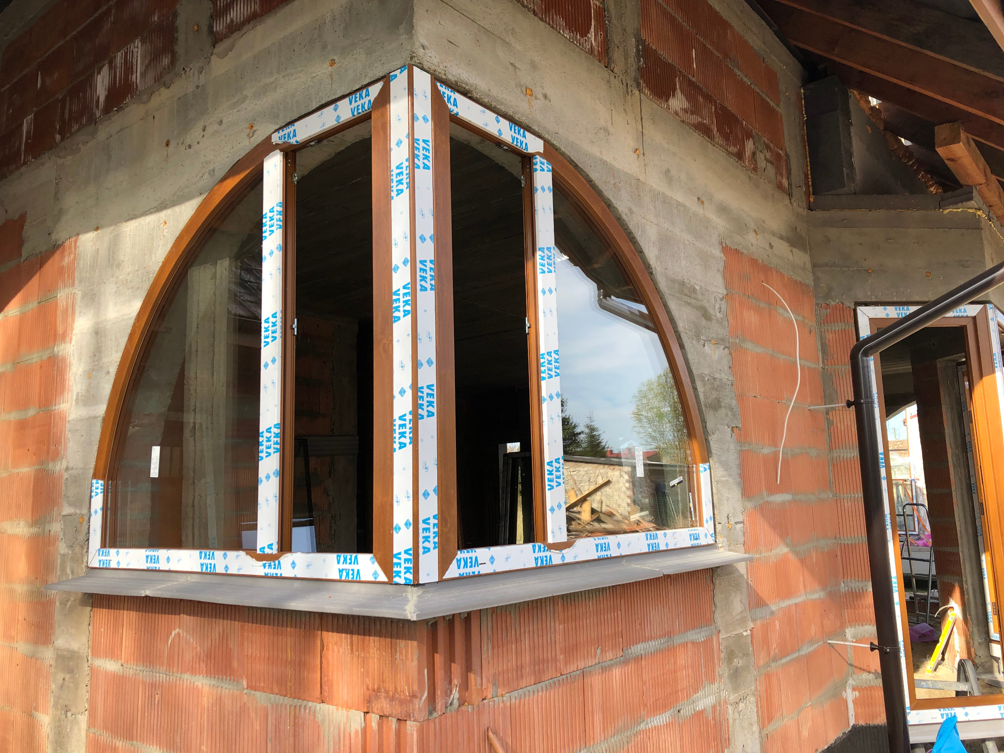 Półokrągłe okno z brązowymi ramami i białymi profilami ochronnymi Veka, zamontowane w surowej ścianie z czerwonej cegły i betonu, widok z zewnątrz.