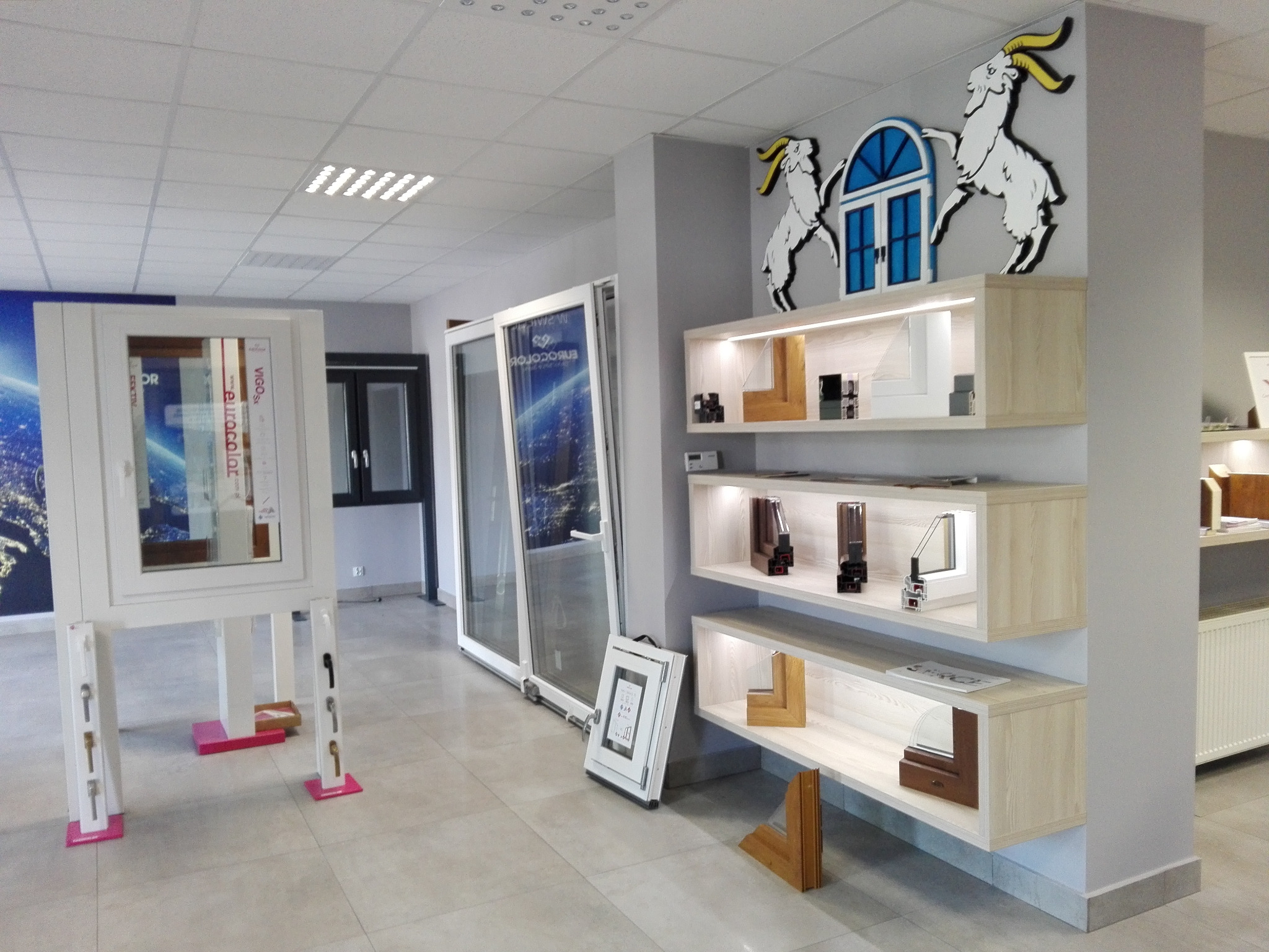 Salon z ekspozycją okien i drzwi, prezentujący różne modele i przekroje profili na półkach, z dekoracyjnymi kozicami na ścianie.