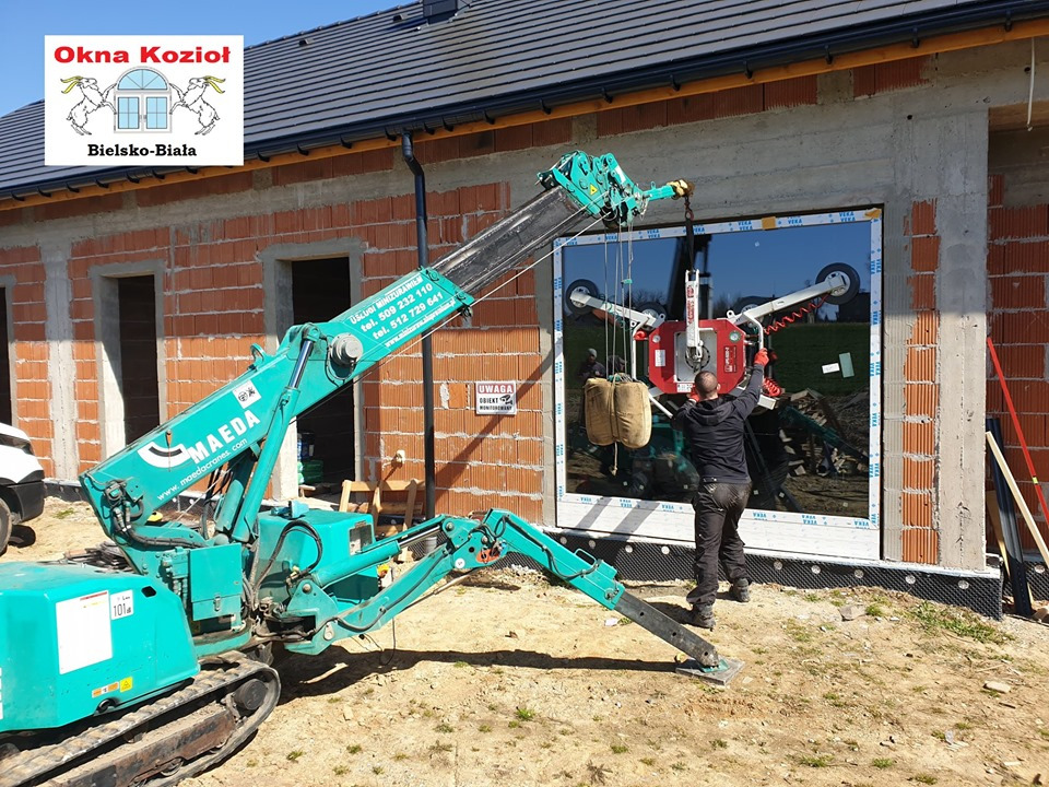 Montaż okna na budowie domu jednorodzinnego za pomocą specjalistycznego miniżurawia Maeda; pracownik w czarnej odzieży kontroluje proces wstawiania szyby z przyssawkami, widoczne logo firmy Okna...