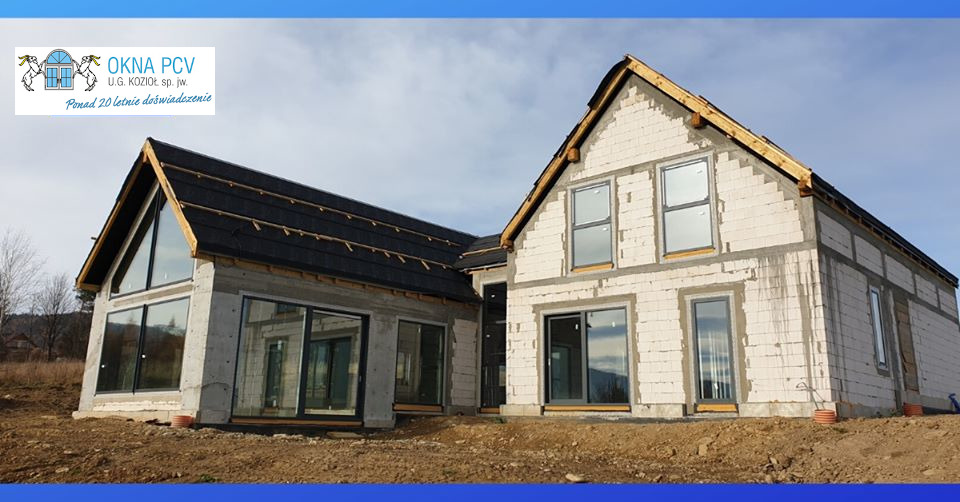 Surowy stan domu w trakcie budowy z zamontowanymi oknami i drzwiami przesuwnymi w szarej ramie, widoczne bloczki betonowe i drewniana konstrukcja dachu, logo firmy na górze zdjęcia.