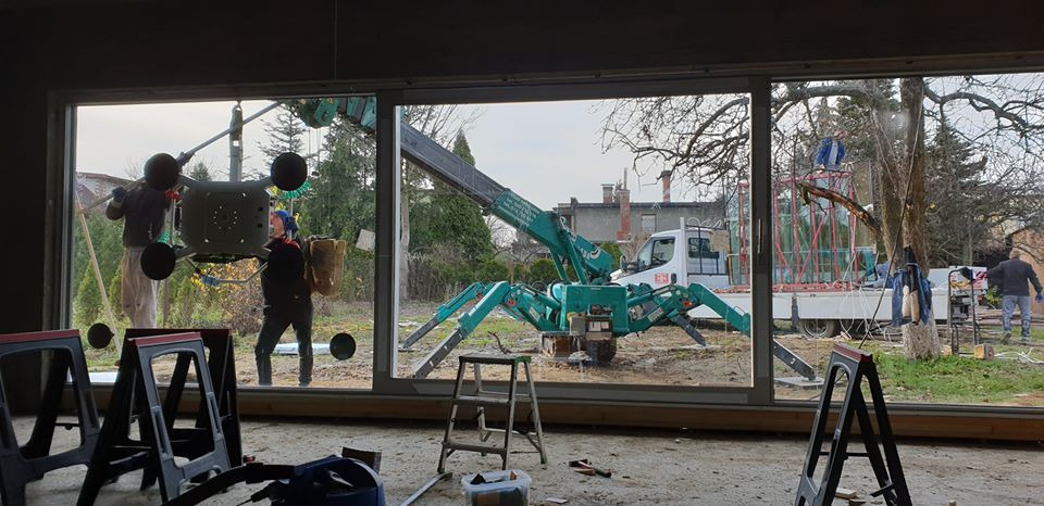 Montaż okna panoramicznego z użyciem przyssawkowej windy i miniżurawia gąsienicowego; widoczni dwaj monterzy, narzędzia oraz transport okien na przyczepie.