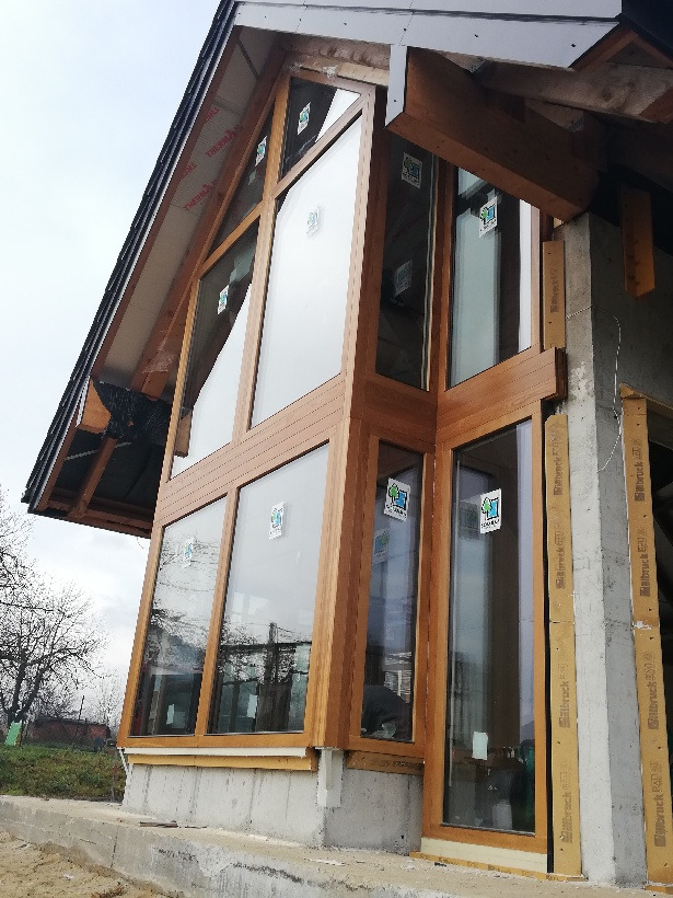 Nowoczesna fasada domu w trakcie budowy z zamontowanymi oknami z drewnopodobnymi ramami, widoczne elementy konstrukcyjne i ocieplenie.