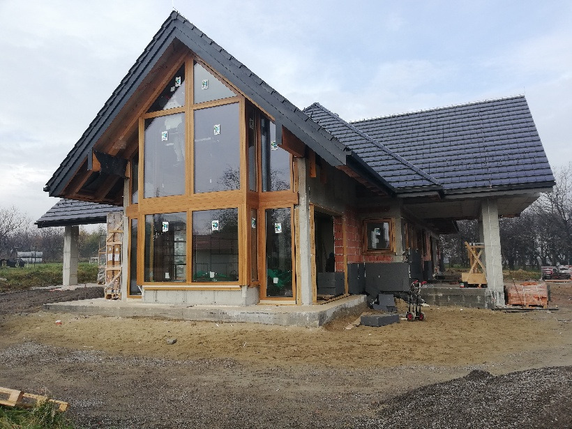 Budynek w trakcie budowy z zamontowanymi dużymi oknami w drewnianych ramach, widoczne materiały budowlane i rusztowania.