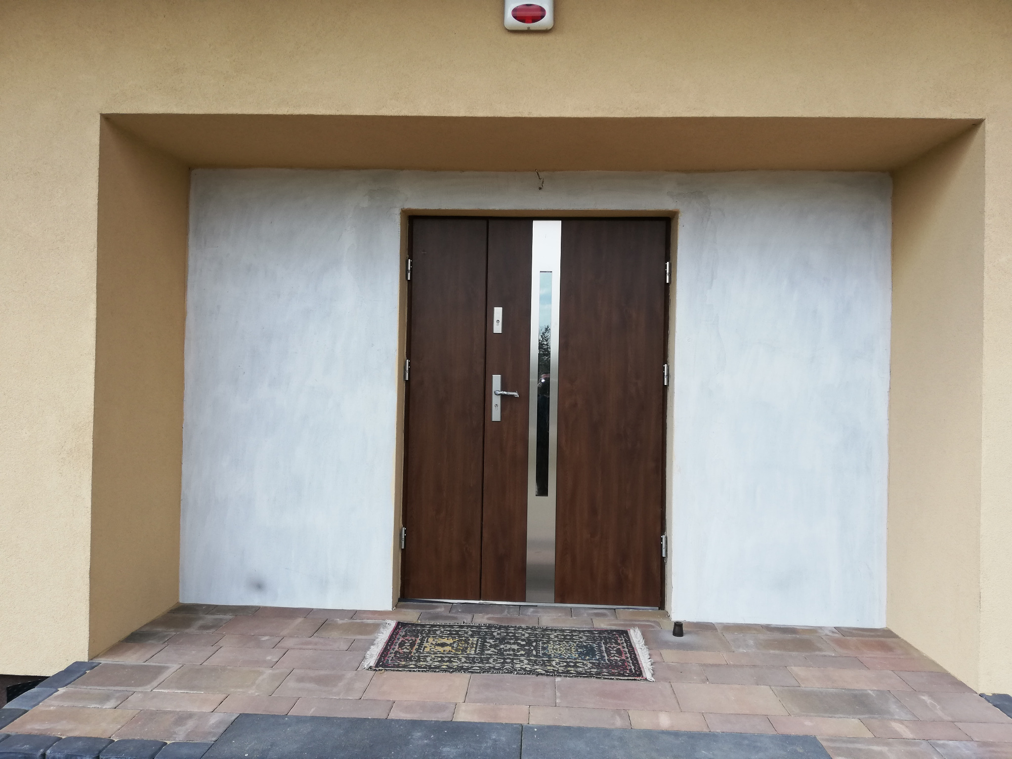 Dwuskrzydłowe drzwi zewnętrzne w kolorze ciemnego drewna z pionowym, wąskim przeszkleniem pośrodku, zamontowane w jasnej wnęce budynku z beżową elewacją, wycieraczka na brązowej kostce brukowej.