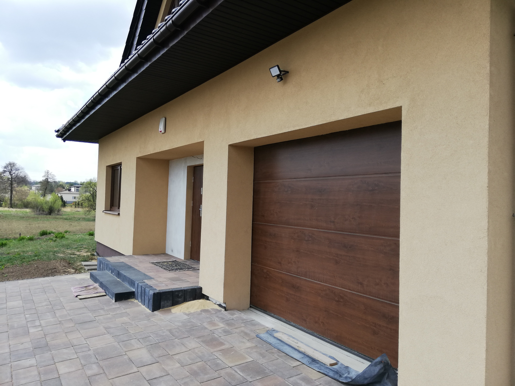Nowoczesna brama garażowa segmentowa w kolorze ciemnego drewna, zamontowana w elewacji domu o beżowym tynku, z widocznymi schodami wejściowymi i kostką brukową.