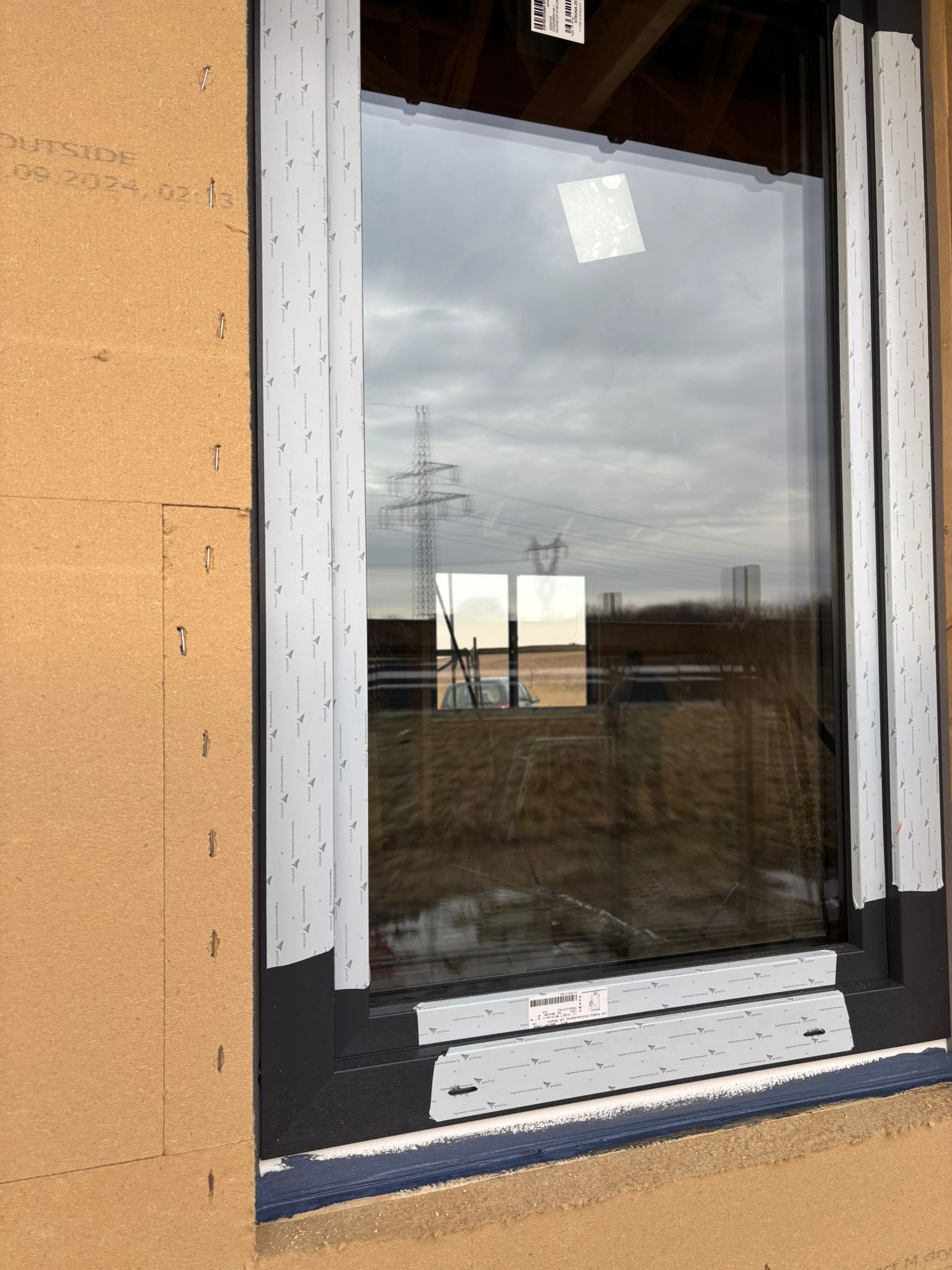 Ciemna rama okienna z widocznymi elementami montażowymi i folią ochronną, elewacja budynku z widocznymi płytami izolacyjnymi i zszywkami, w tle słup wysokiego napięcia i pochmurne niebo.