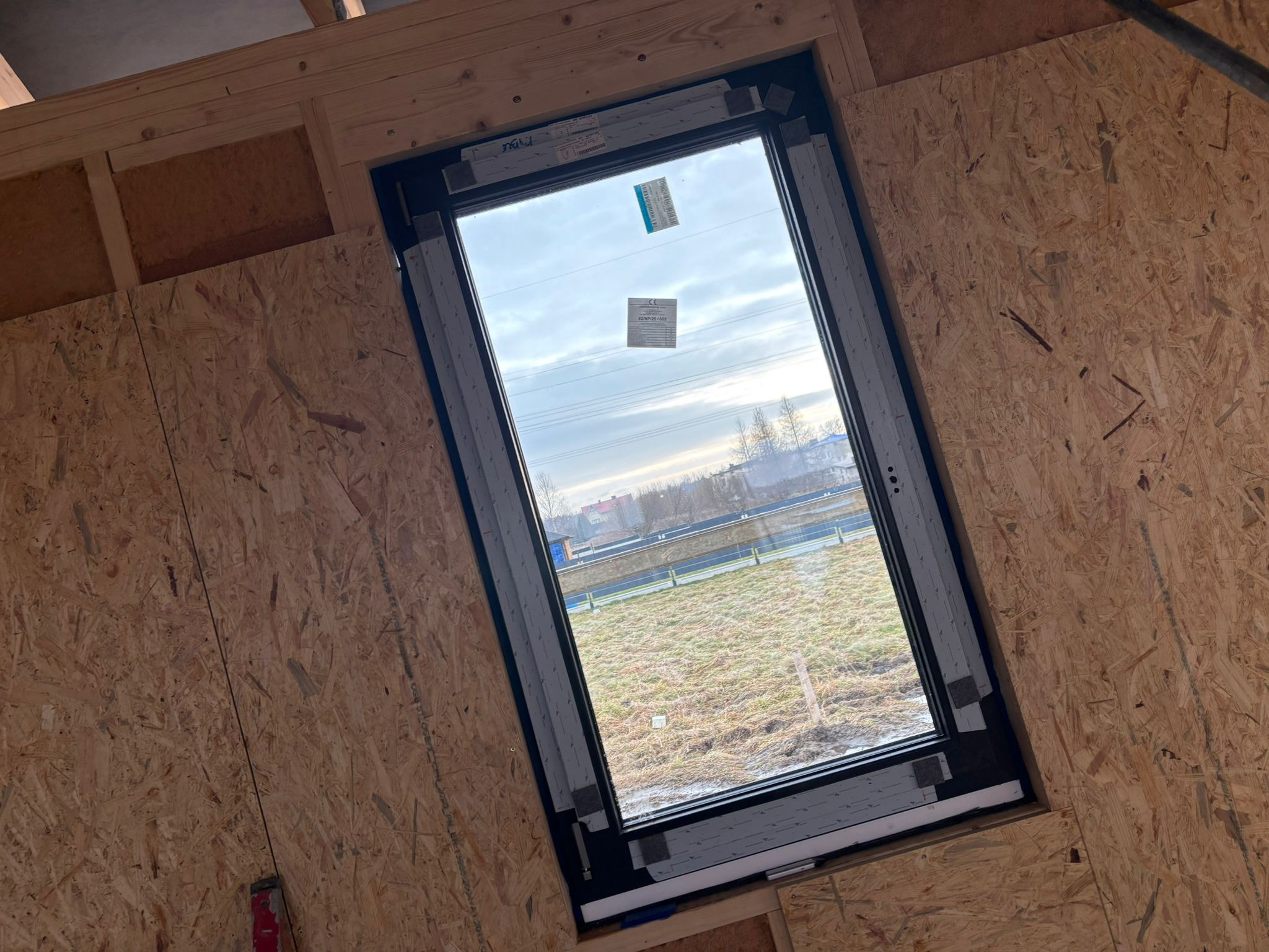 Montaż okna dachowego w konstrukcji szkieletowej, widoczne uszczelki i naklejki producenta na szybie, tło stanowią pola i zabudowania.