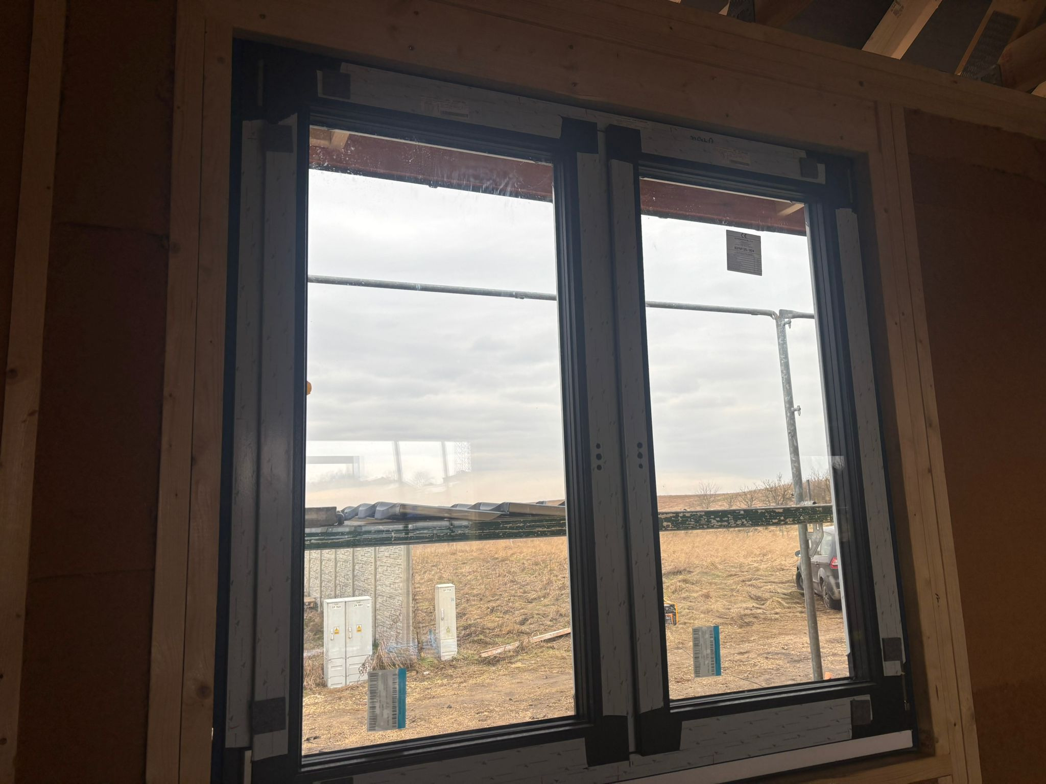 Montaż okna dwukomorowego w surowym stanie; widoczne drewniane elementy konstrukcyjne budynku, folia ochronna na szybach, oraz krajobraz za oknem z polami i skrzynkami elektrycznymi.