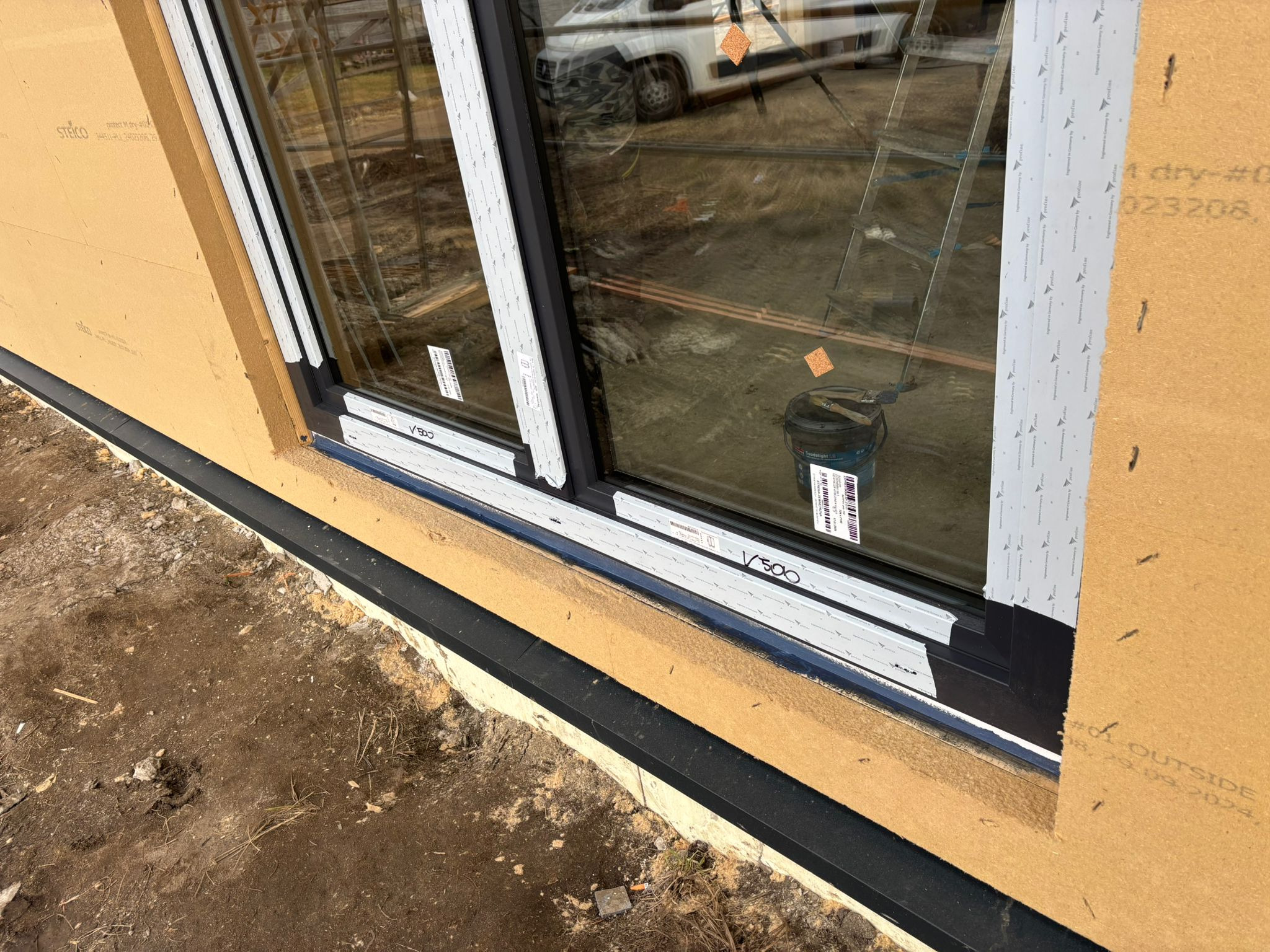 Montaż okna PCV z czarną ramą w surowym stanie, widoczna warstwa izolacyjna na elewacji, odbicie samochodu w szybie, wiadro z farbą wewnątrz budynku.