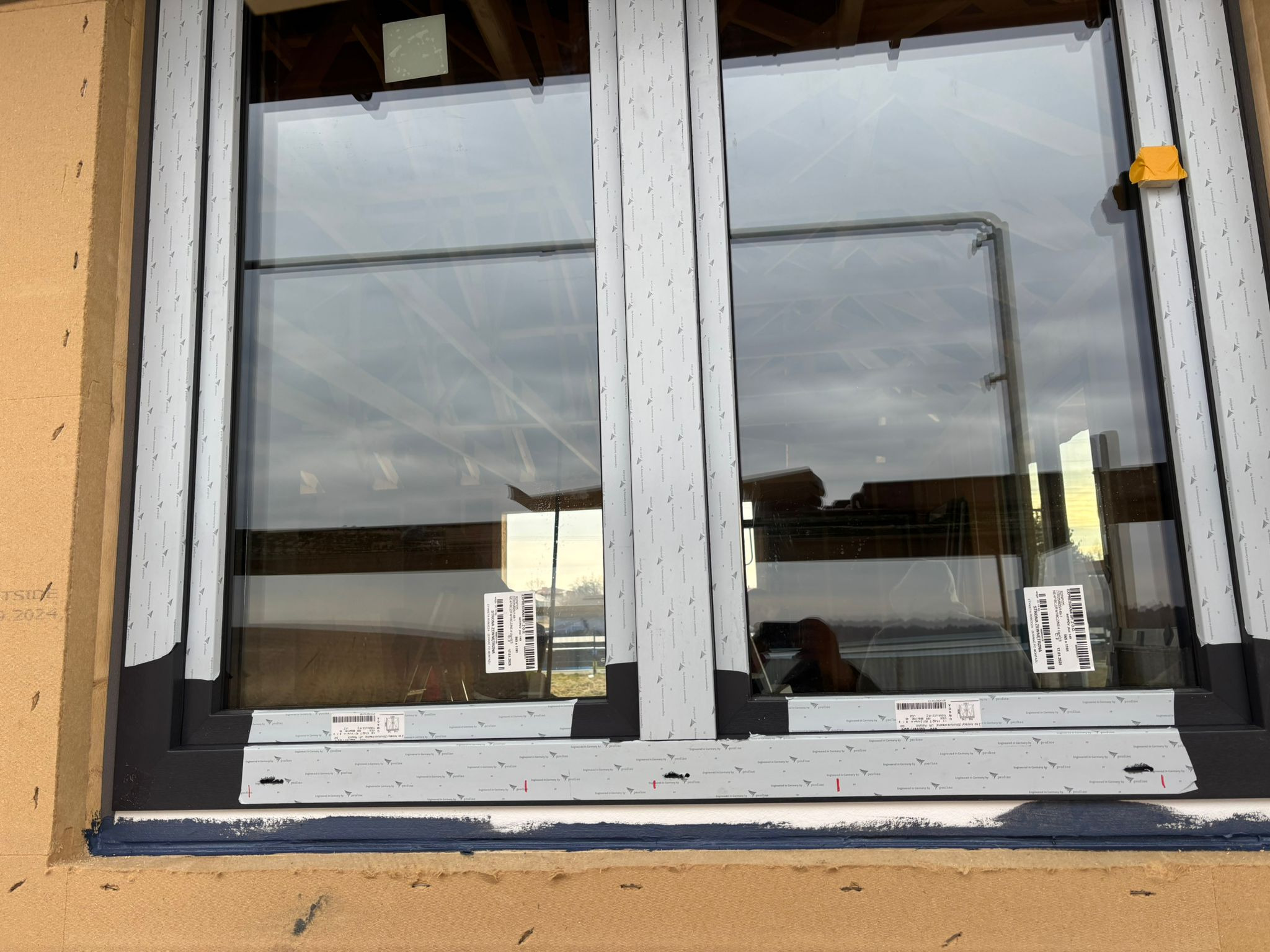 Montaż dwuskrzydłowego okna z czarną ramą, zabezpieczonego taśmą montażową z nadrukiem, widoczne etykiety z kodami kreskowymi na szybach, oraz żółta taśma zabezpieczająca na górnym rogu ramy.